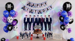 Mise en scène d'une table d'anniversaire décorée avec le kit festif sur le thème de BTS. Au mur, une affiche des 7 chanteurs et une guirlande 'Happy Birthday' surplombent un gâteau au chocolat. De part et d'autre, de généreux bouquets de ballons bleus, violets, noirs et blancs avec le logo du groupe. Sur la table sont disposés des gobelets rouges et les petites décorations cartonnées à l'effigie des membres du groupe K-Pop.