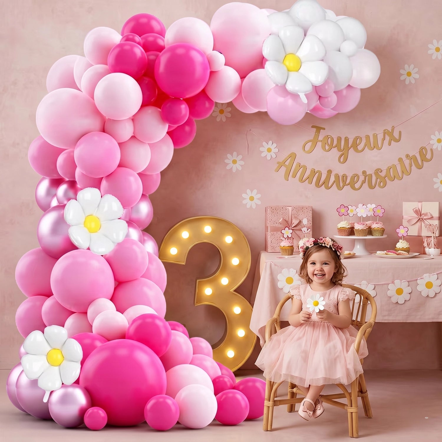 Arche de ballons fuchsia, rose et marguerite décorant un coin d'anniversaire d'enfant pour les 3 ans. Elle encadre un grand numéro 3 lumineux et une table de desserts avec cupcakes. Une petite fille en robe de fête est assise sur une chaise en rotin."