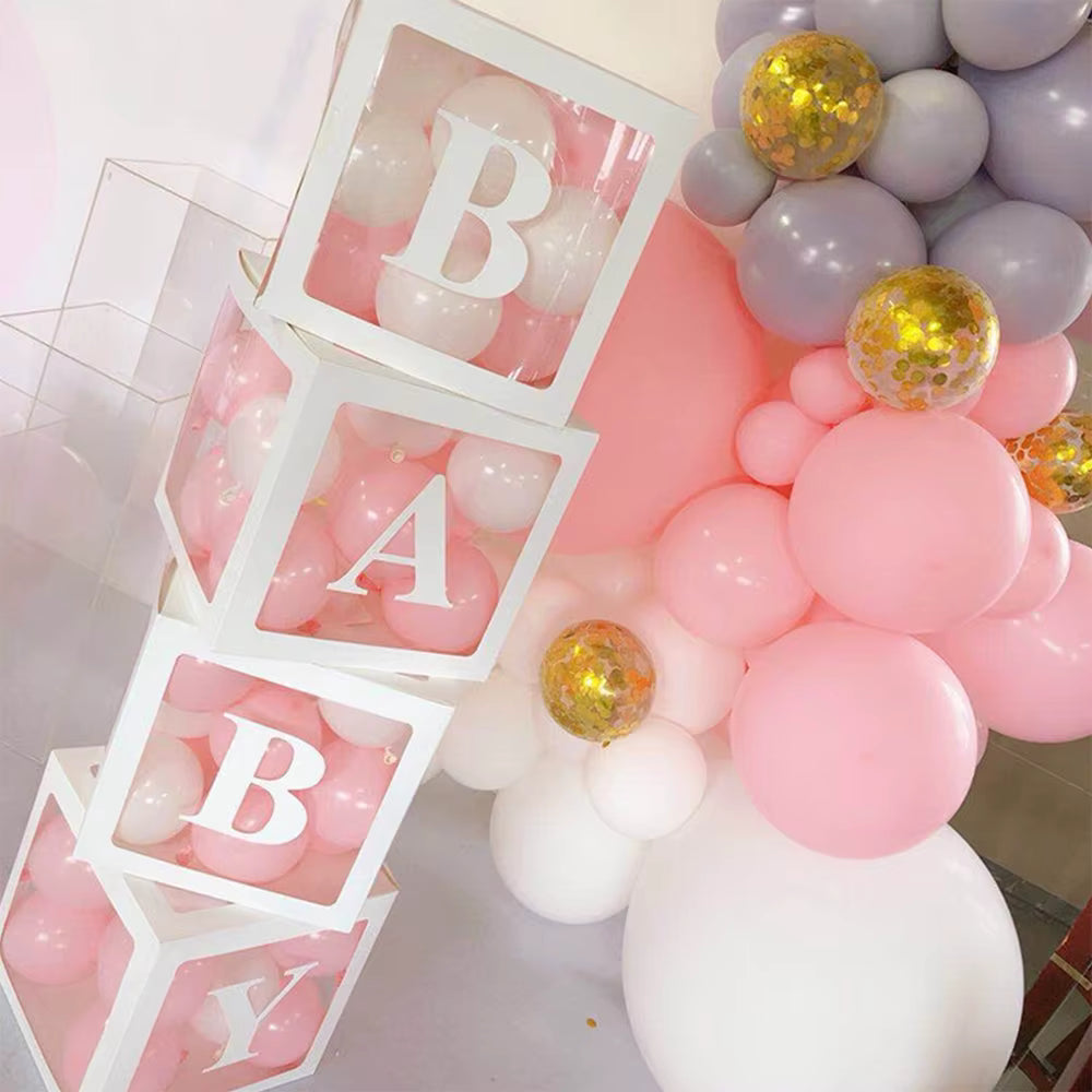 Vue en gros plan d'une pile verticale de quatre boîtes à ballons transparentes formant 'BABY', remplies de petits ballons blancs et roses. Elles sont disposées à côté d'une grappe de ballons de fête roses, gris et confettis dorés.