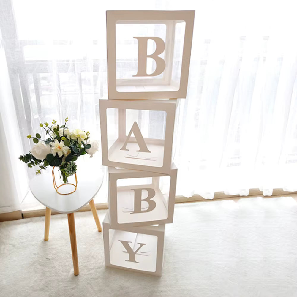 Pile de quatre boîtes de décoration pour bébé transparentes et vides, alignées verticalement pour former le mot 'BABY' avec des lettres blanches découpées. Le décor est un intérieur lumineux avec une petite table scandinave et un vase de fleurs blanches et eucalyptus à gauche.