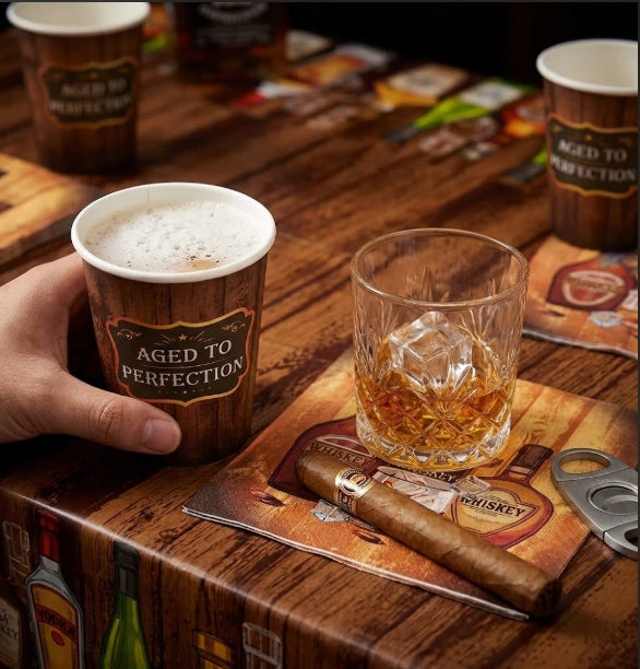 Gros plan macro sur une table de fête avec un gobelet en carton "Aged to Perfection" rempli de bière, un verre de whisky ambré avec glaçons posé sur une serviette illustrée, et un cigare avec son coupe-cigare.
